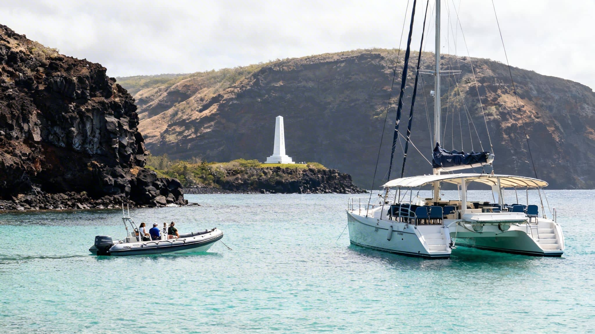 A catamaran and an inflatable boat with people in clear blue water, with a white monument on a rocky island and hills.