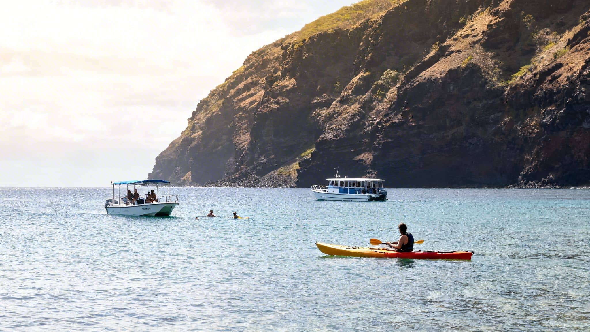 People kayaking, swimming, and boating in Kealakekua Bay with a scenic cliff.