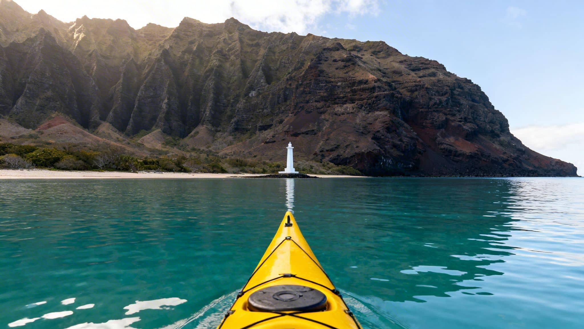 A yellow kayak paddles towards a white lighthouse on a tropical beach with towering mountains.