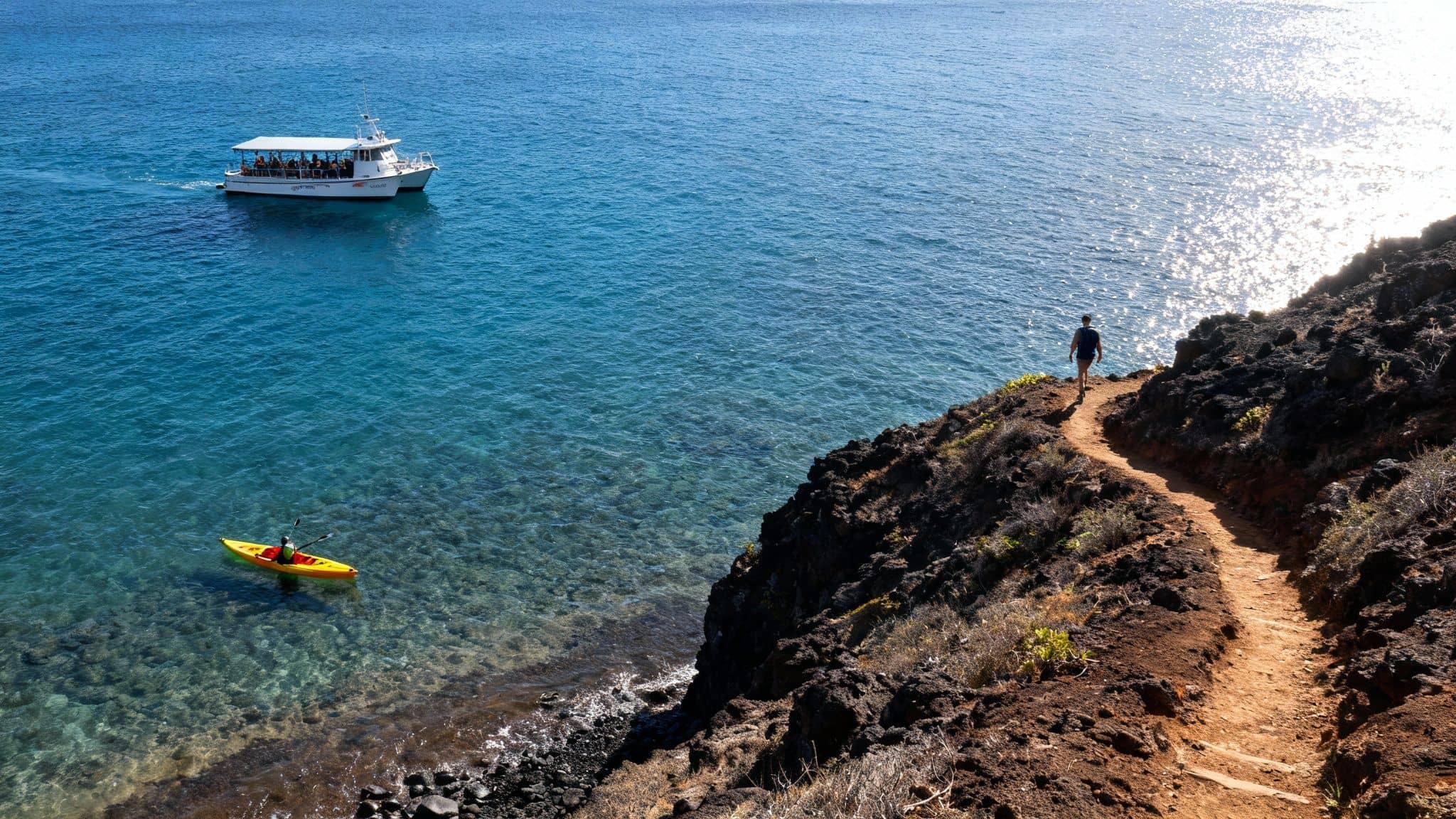A man hikes a rugged coastal trail overlooking a boat and a kayaker in clear blue ocean.