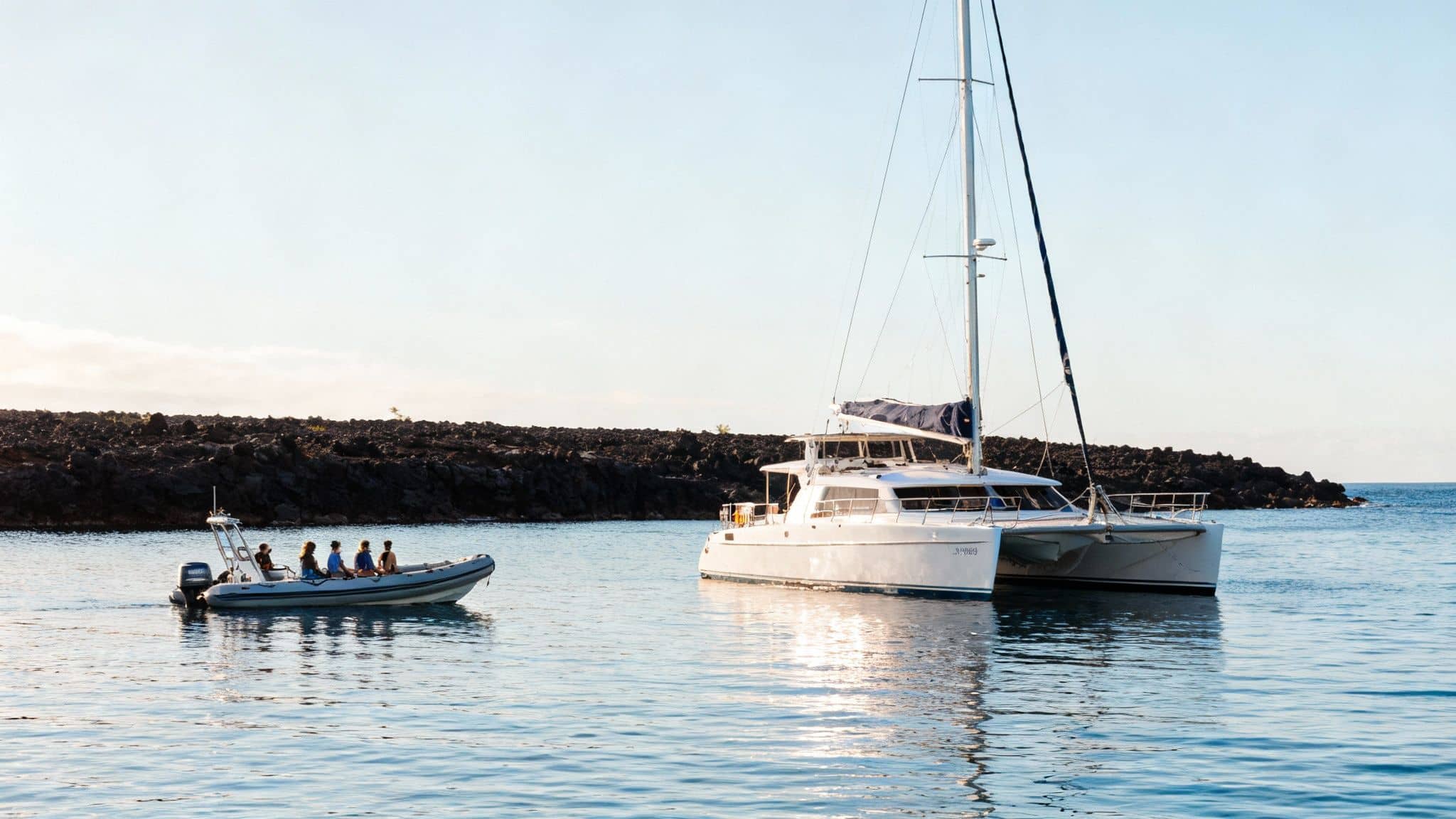 A large white catamaran sailboat and a dinghy with people on calm water near a rocky island.