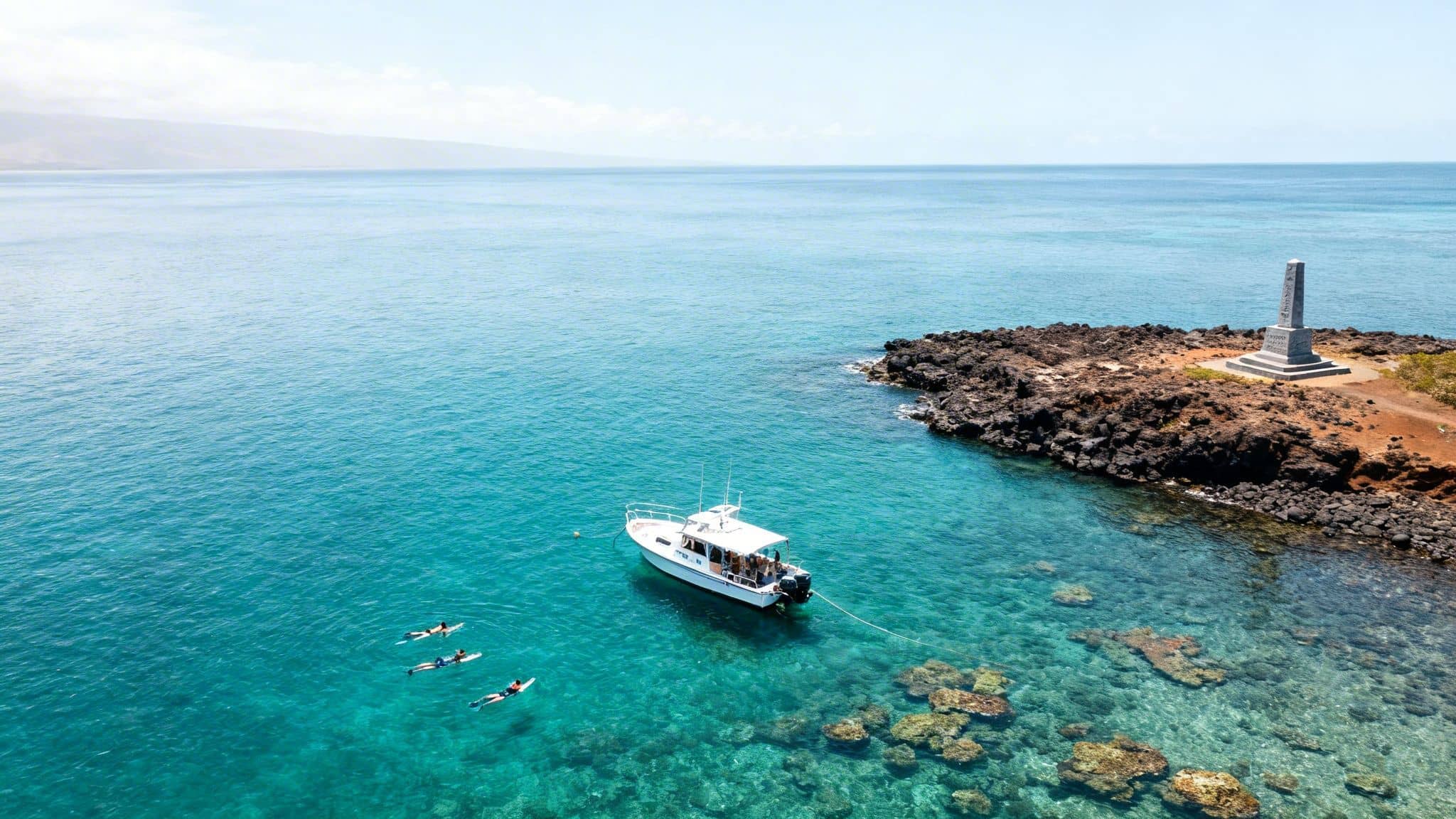 Aerial view of a boat, snorkelers, and a monument in clear turquoise water near a rocky shore.