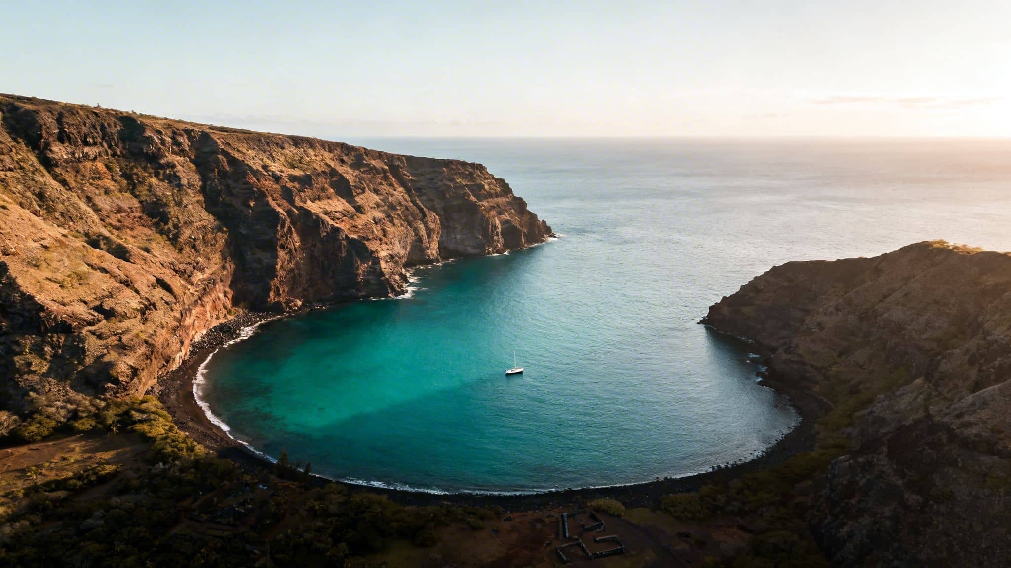 Aerial view of a secluded bay with turquoise water, rugged cliffs, and a sailboat at anchor.