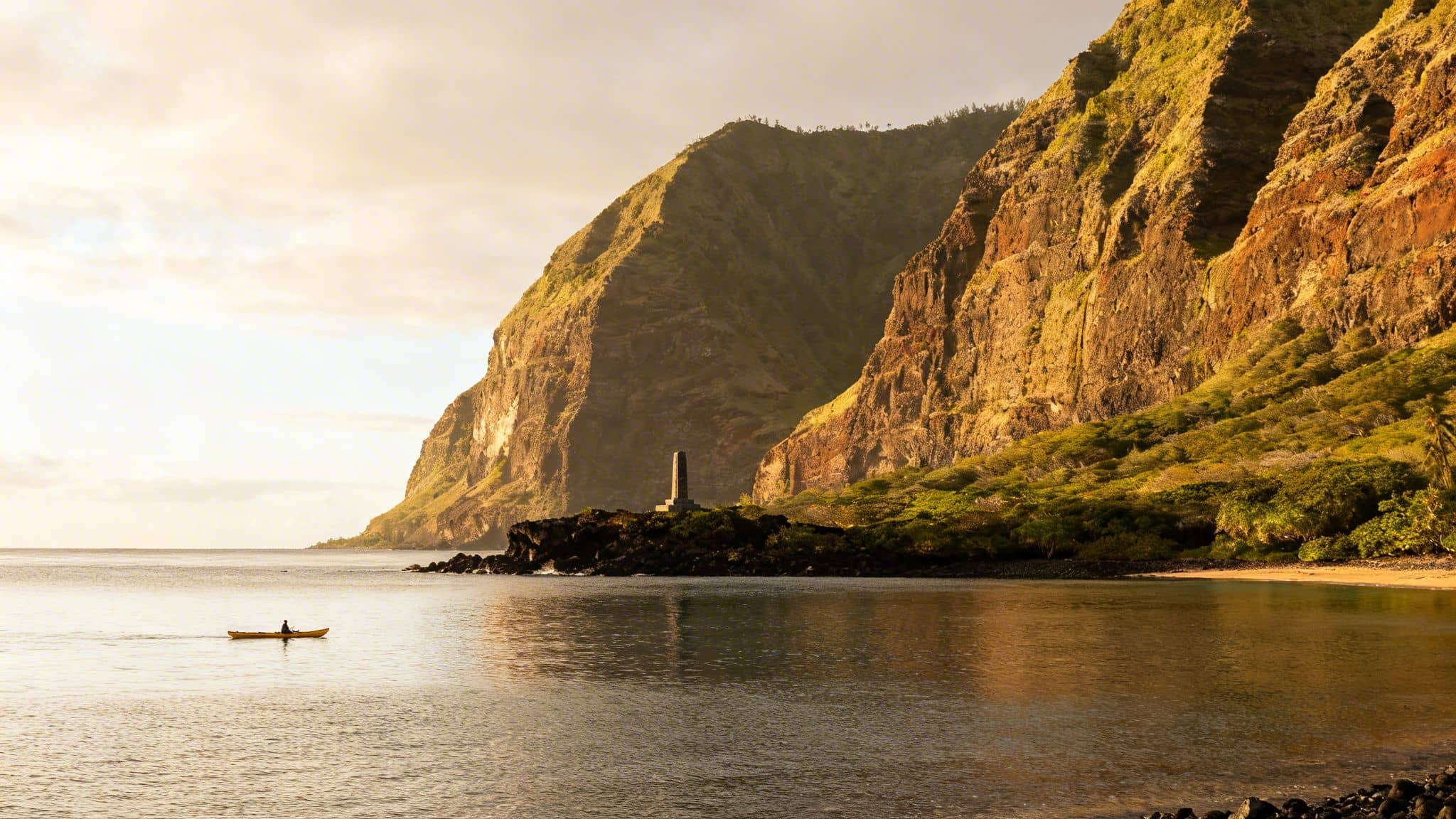 A person kayaks in golden water near majestic coastal cliffs and a historic monument at sunrise.