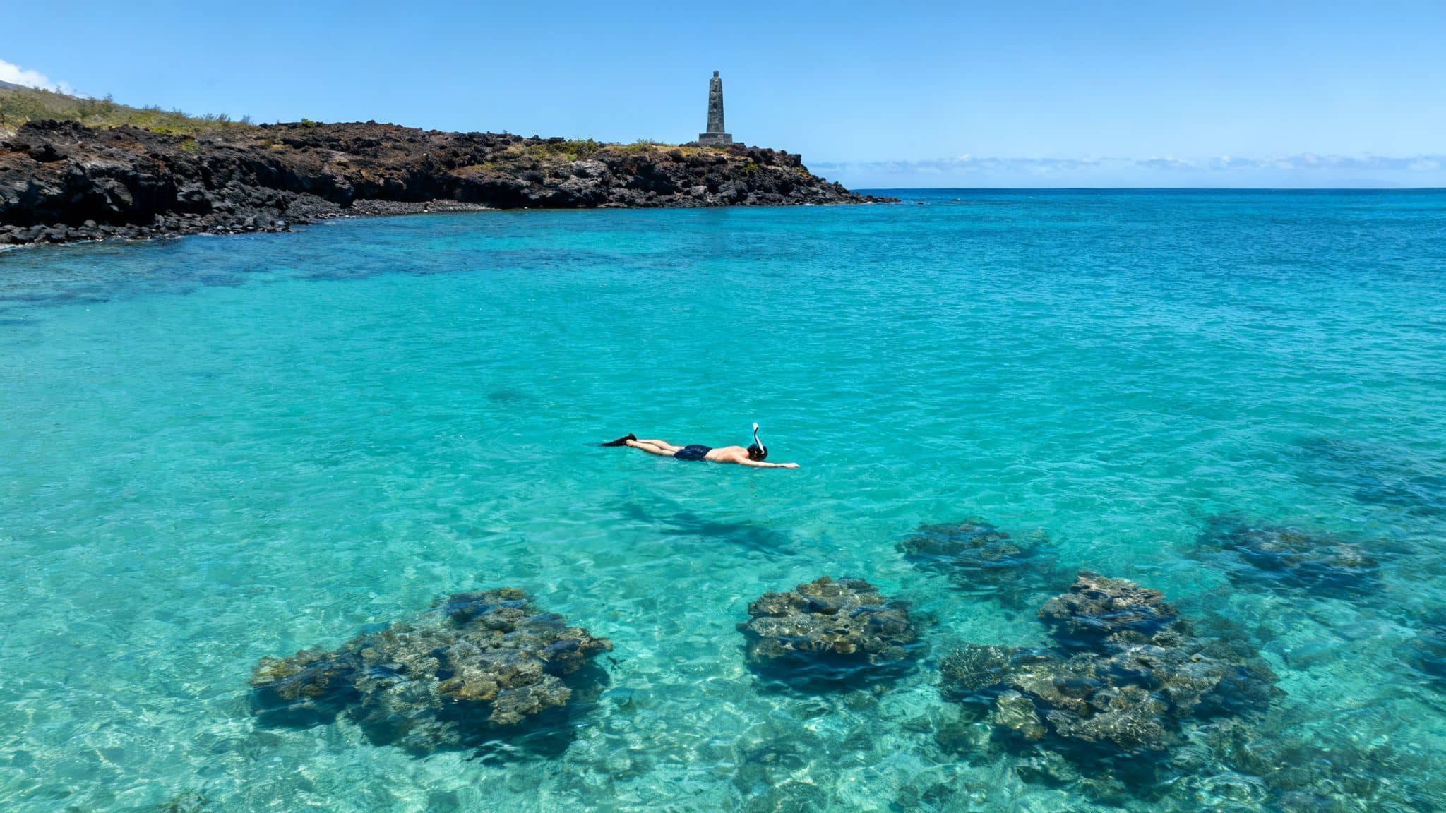 A person snorkeling face down in crystal-clear turquoise water near a rocky Hawaiian coast with a monument.