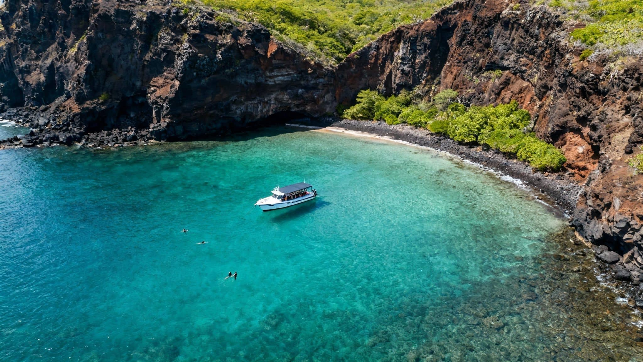 Aerial view of a boat in Kealakekua Bay, Hawaii, with snorkelers near volcanic cliffs and green foliage.