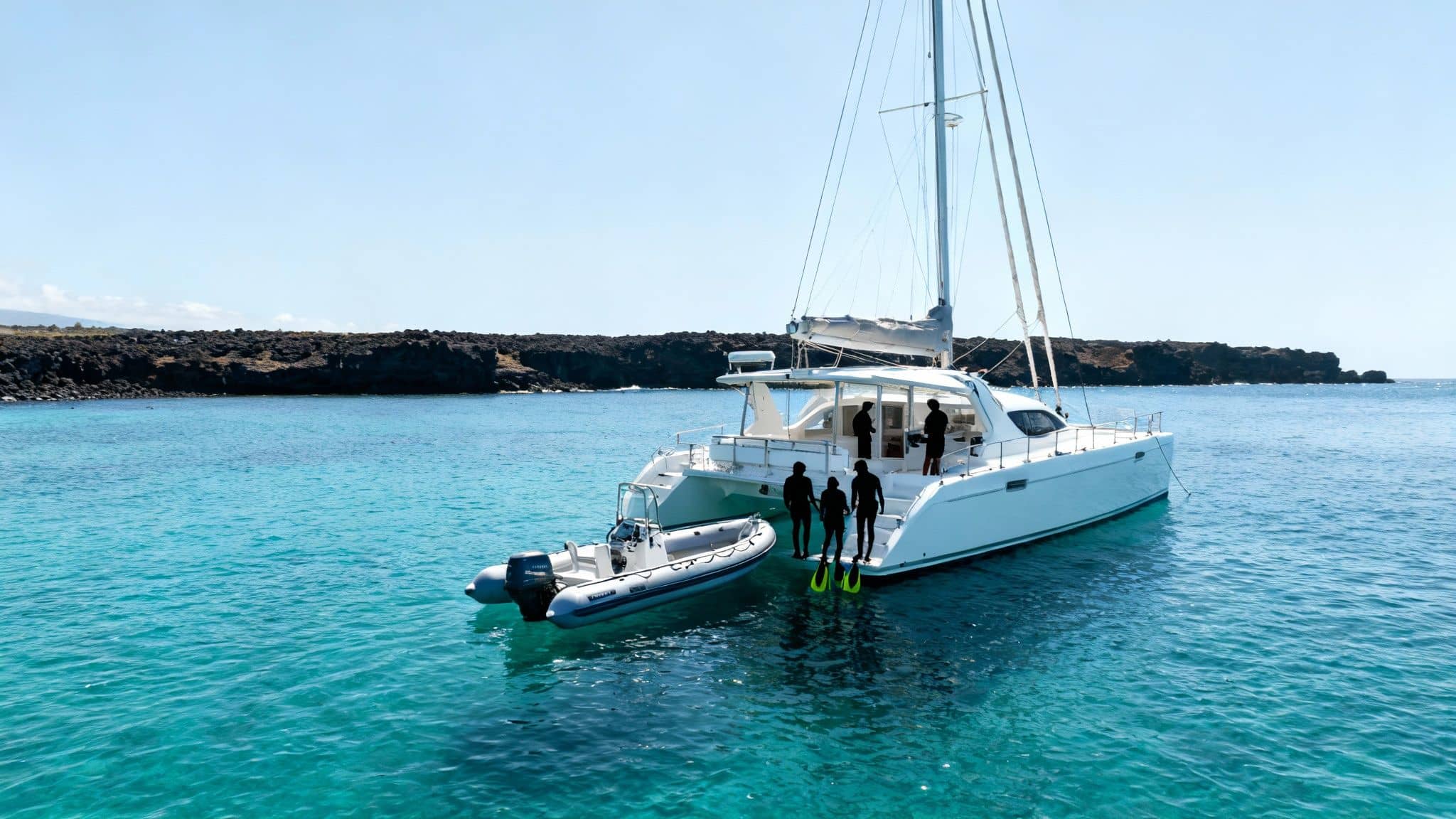 People on a white catamaran and small boat in clear blue water near a rocky coast, preparing to snorkel.