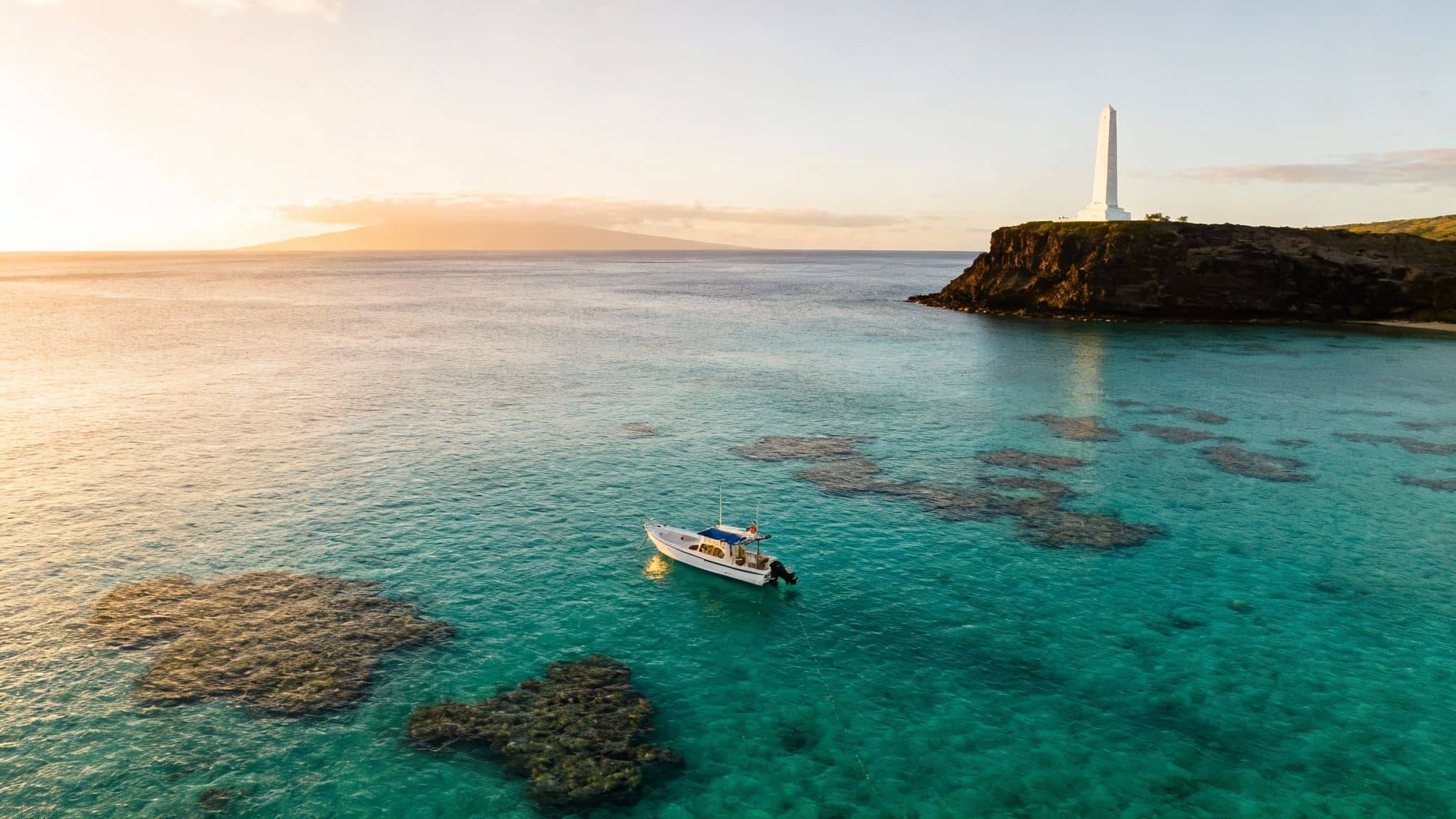 Aerial view of a boat on clear turquoise water with vibrant coral reefs, a cliff monument, and distant island.