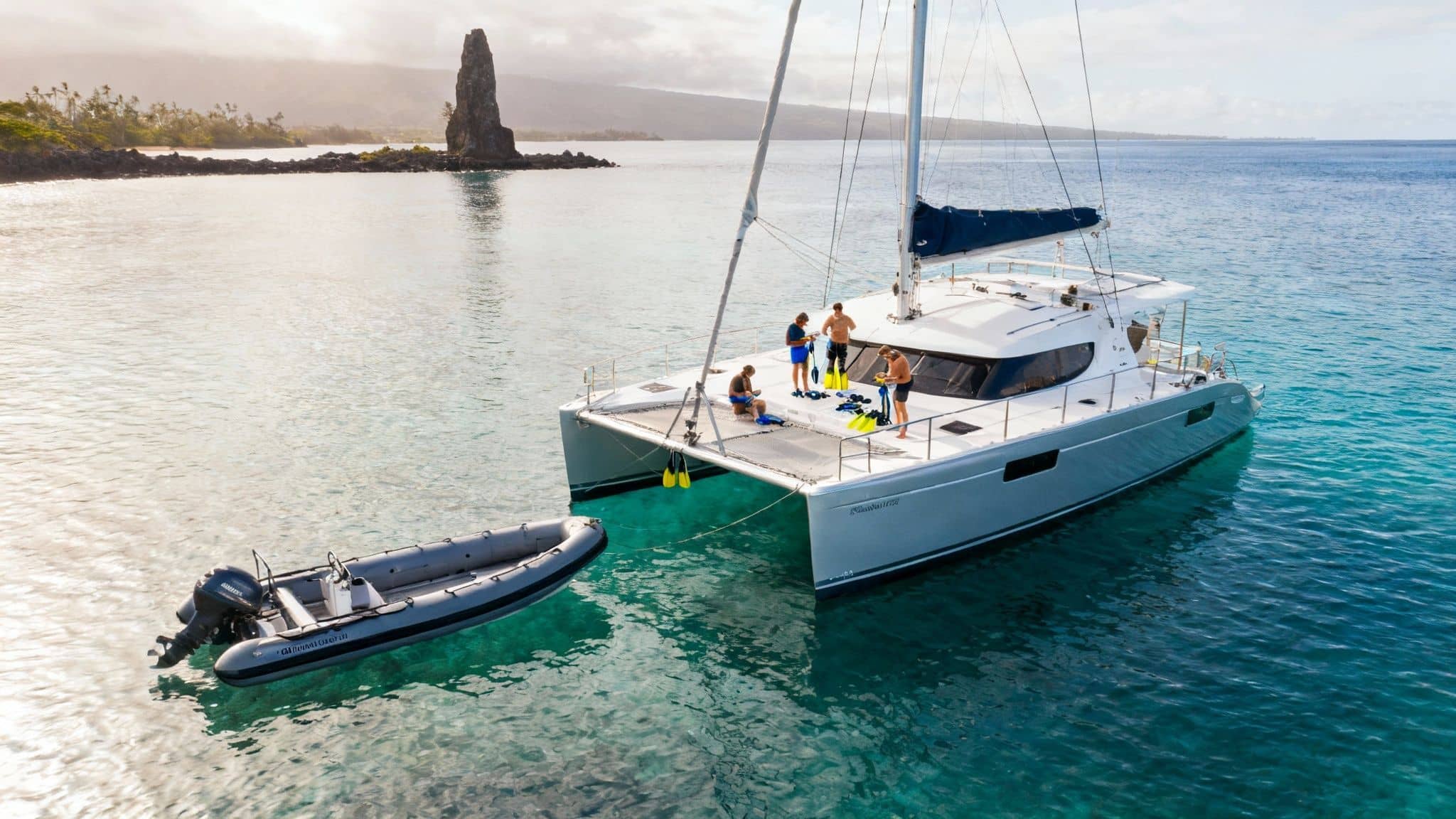 People prepare to snorkel on a catamaran anchored in clear tropical waters, a dinghy floats nearby.
