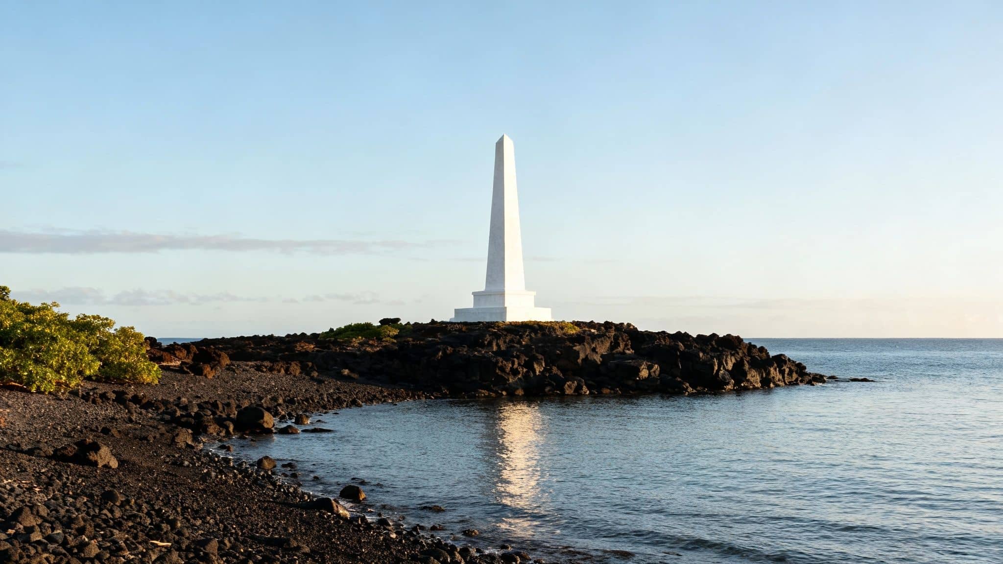 A white obelisk, the Captain Cook Monument, standing on the shore of Kealakekua Bay with lush green cliffs in the background and clear blue water in the foreground.