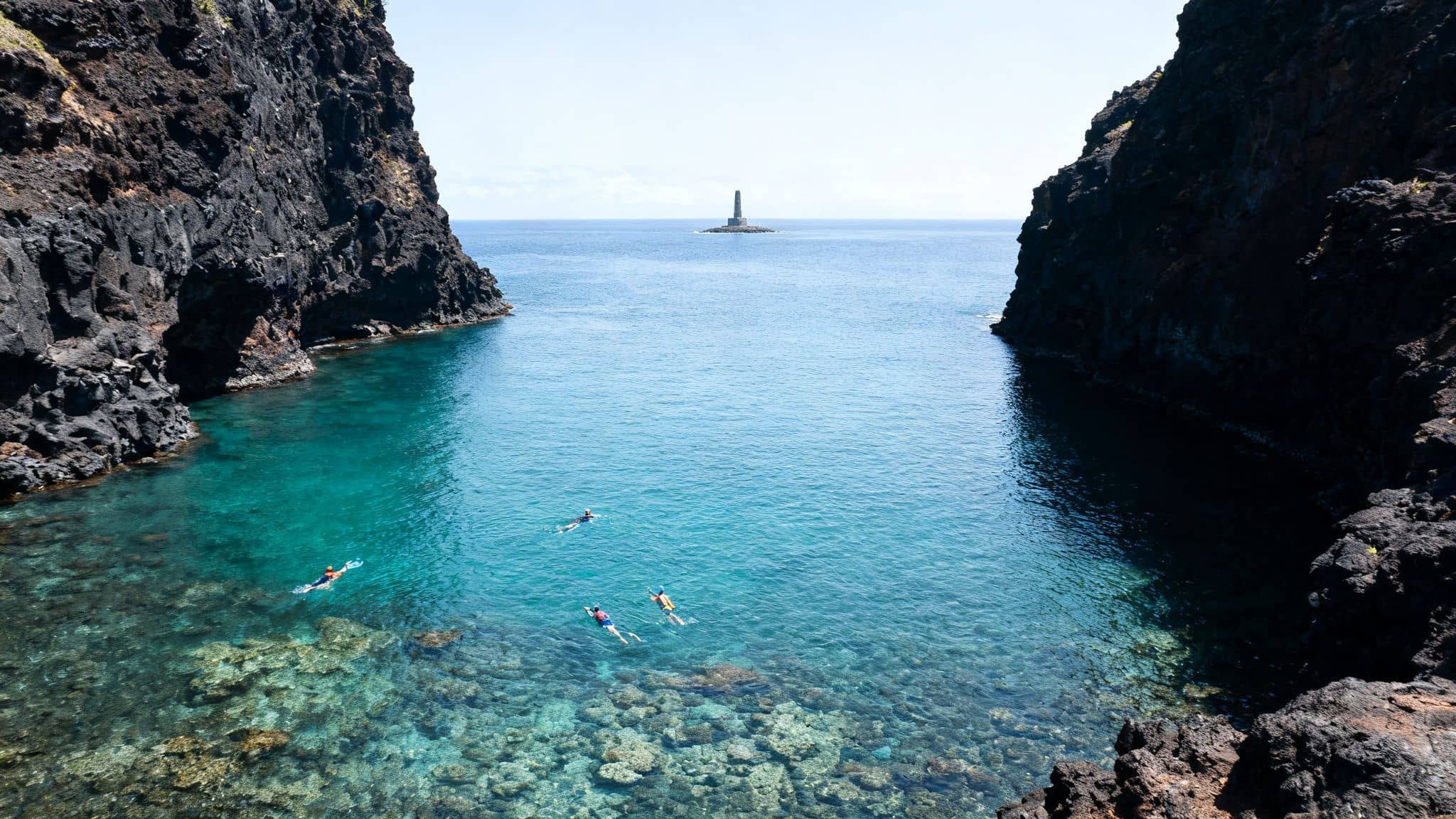 A high-angle shot of people snorkeling in a clear blue cove between dark cliffs, with a distant lighthouse.