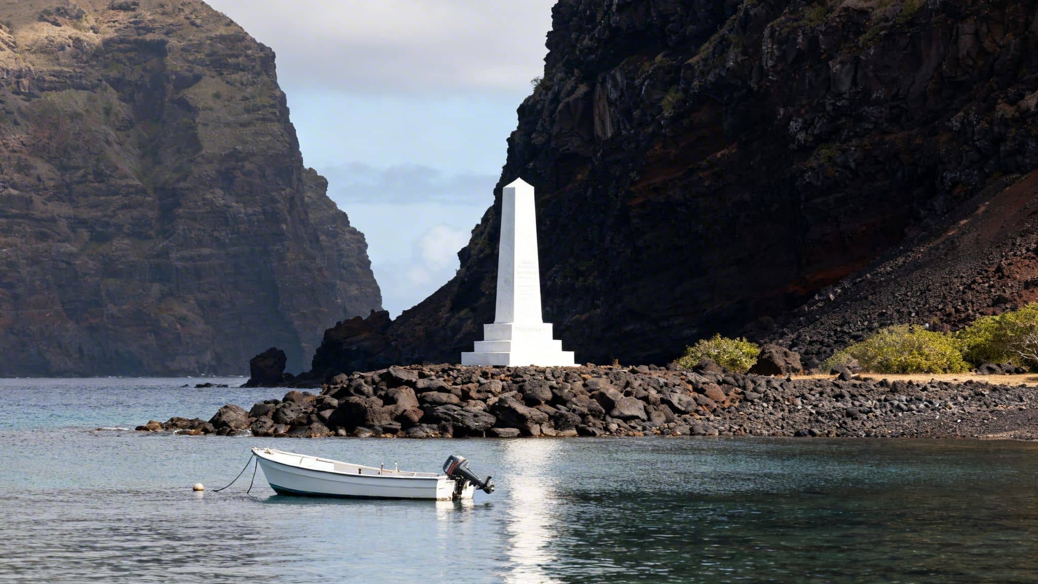 White obelisk monument on volcanic rocks, a small boat in a tranquil bay with towering dark cliffs.