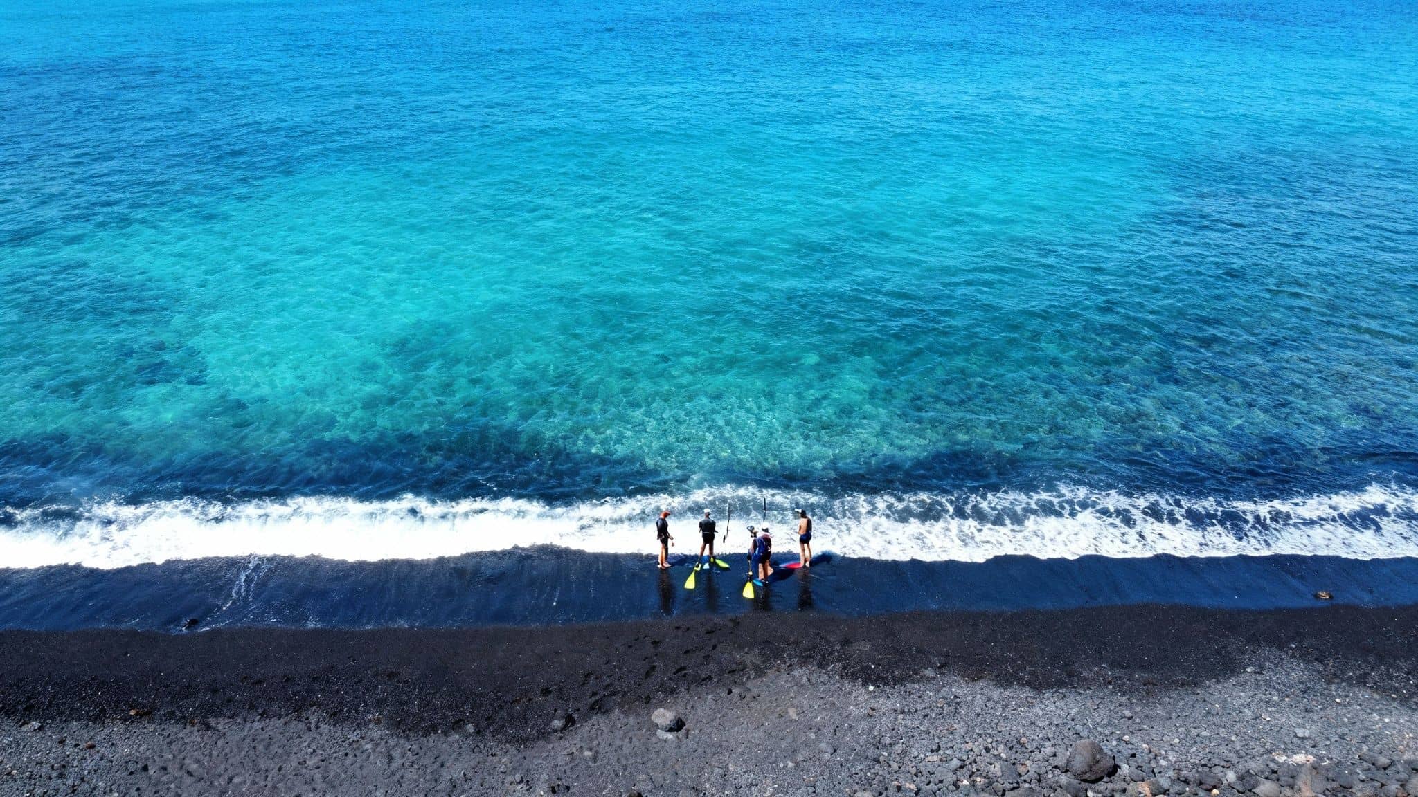 Aerial view of four people with snorkeling gear on a black sand beach next to clear blue ocean water.