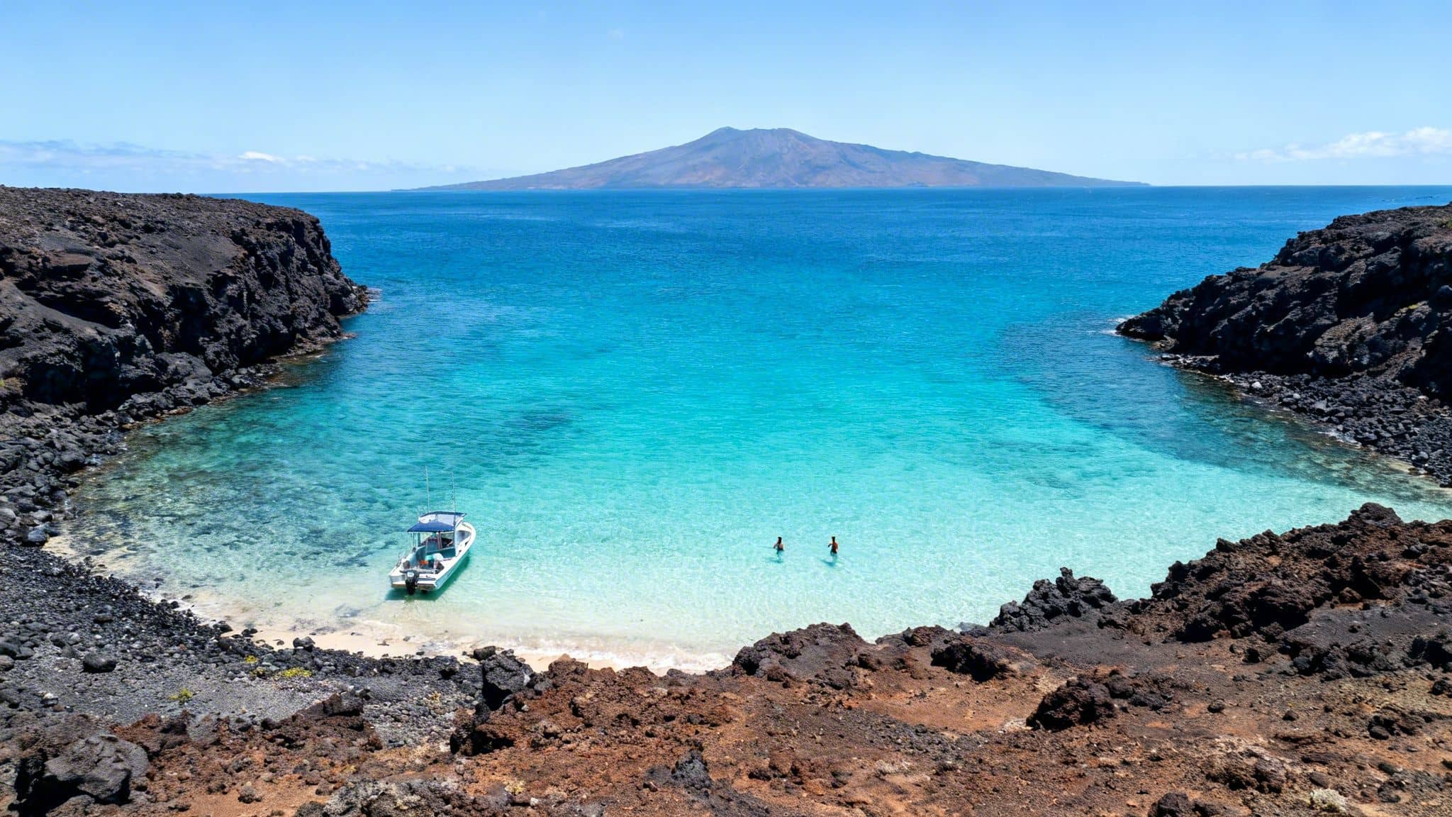 A stunning turquoise bay with a boat and two swimmers, surrounded by dark volcanic rocks, with a distant island under a clear sky.