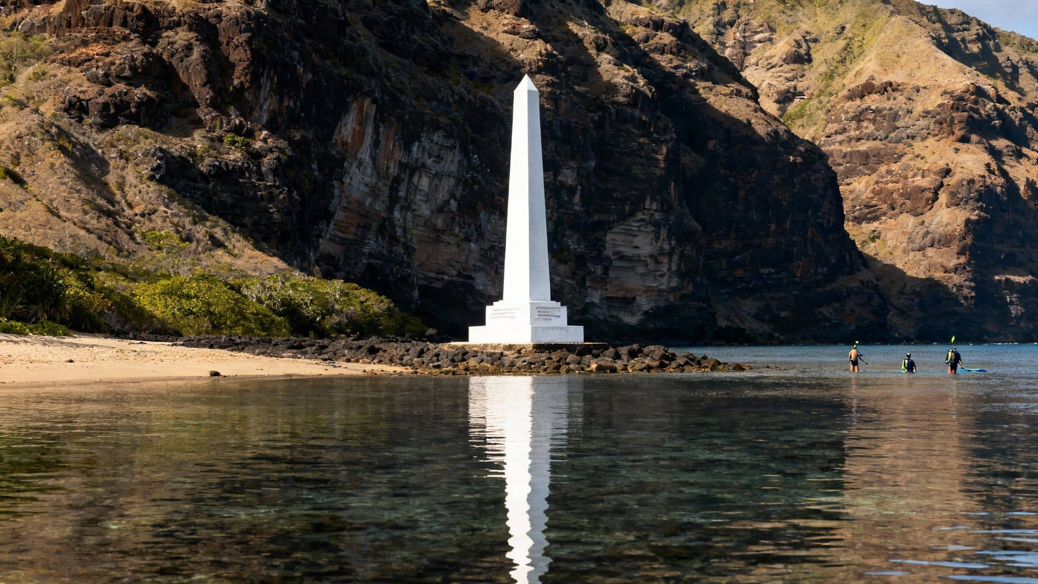 Scenic view of a white obelisk on a rocky beach with snorkelers in clear ocean water.