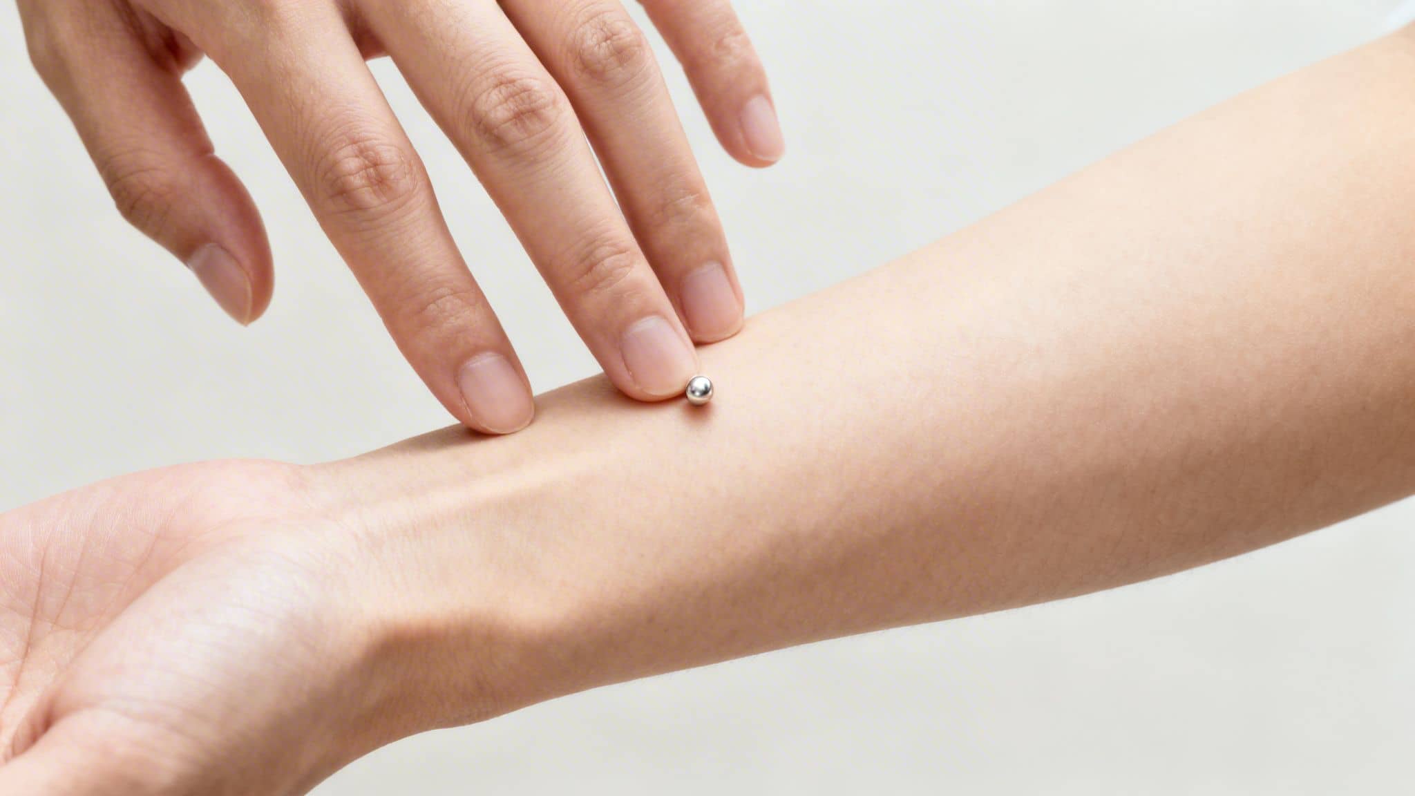A hand's fingers gently touch a small silver bead on another person's pale forearm, likely for acupressure.