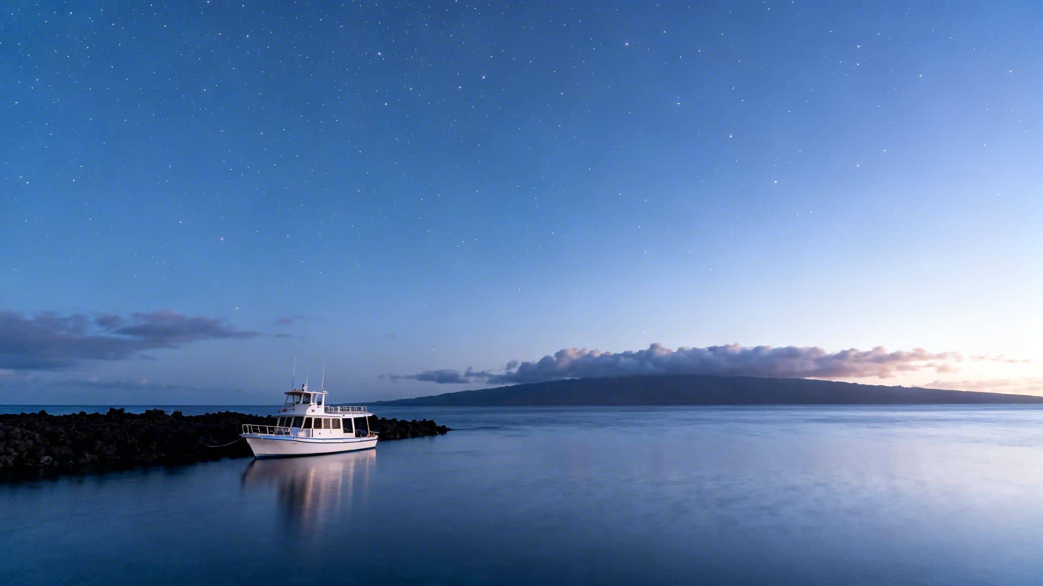 White boat moored by volcanic rocks at night with a starry sky over calm ocean and distant land.
