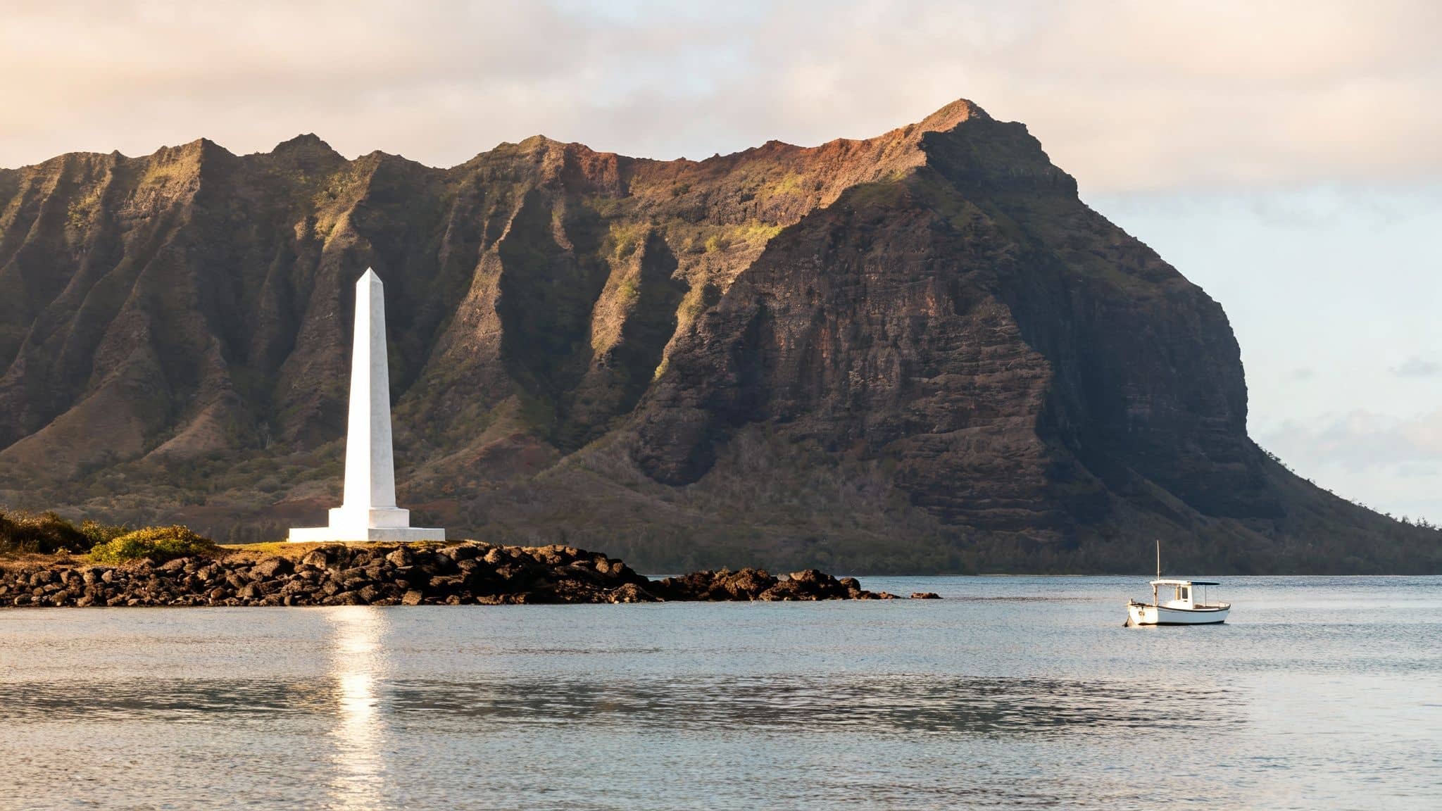 A tall white obelisk stands on a rocky shore, with majestic mountains and a small boat at sunset.