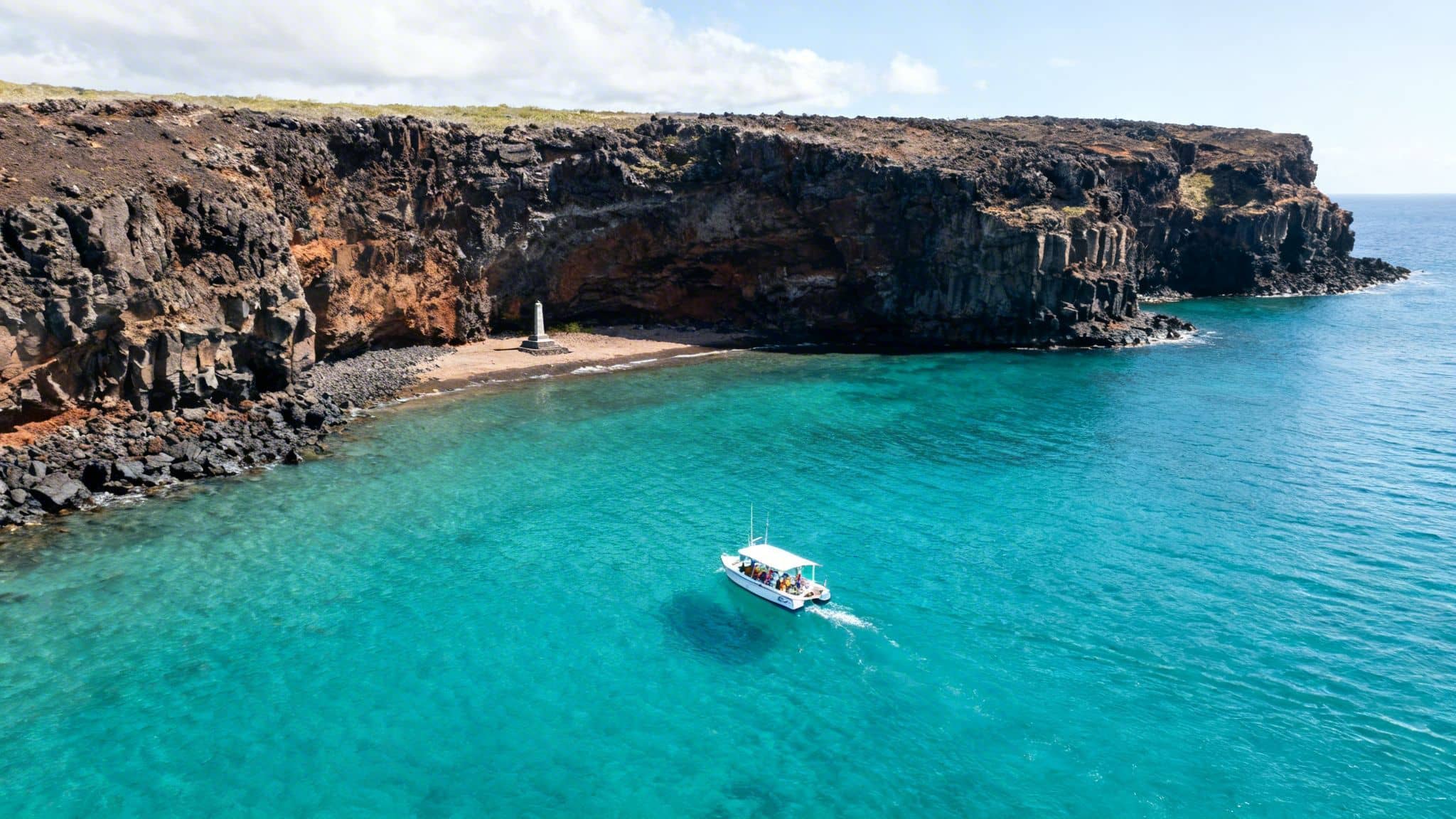 A white tour boat with people on clear turquoise water near rocky volcanic cliffs and a monument.