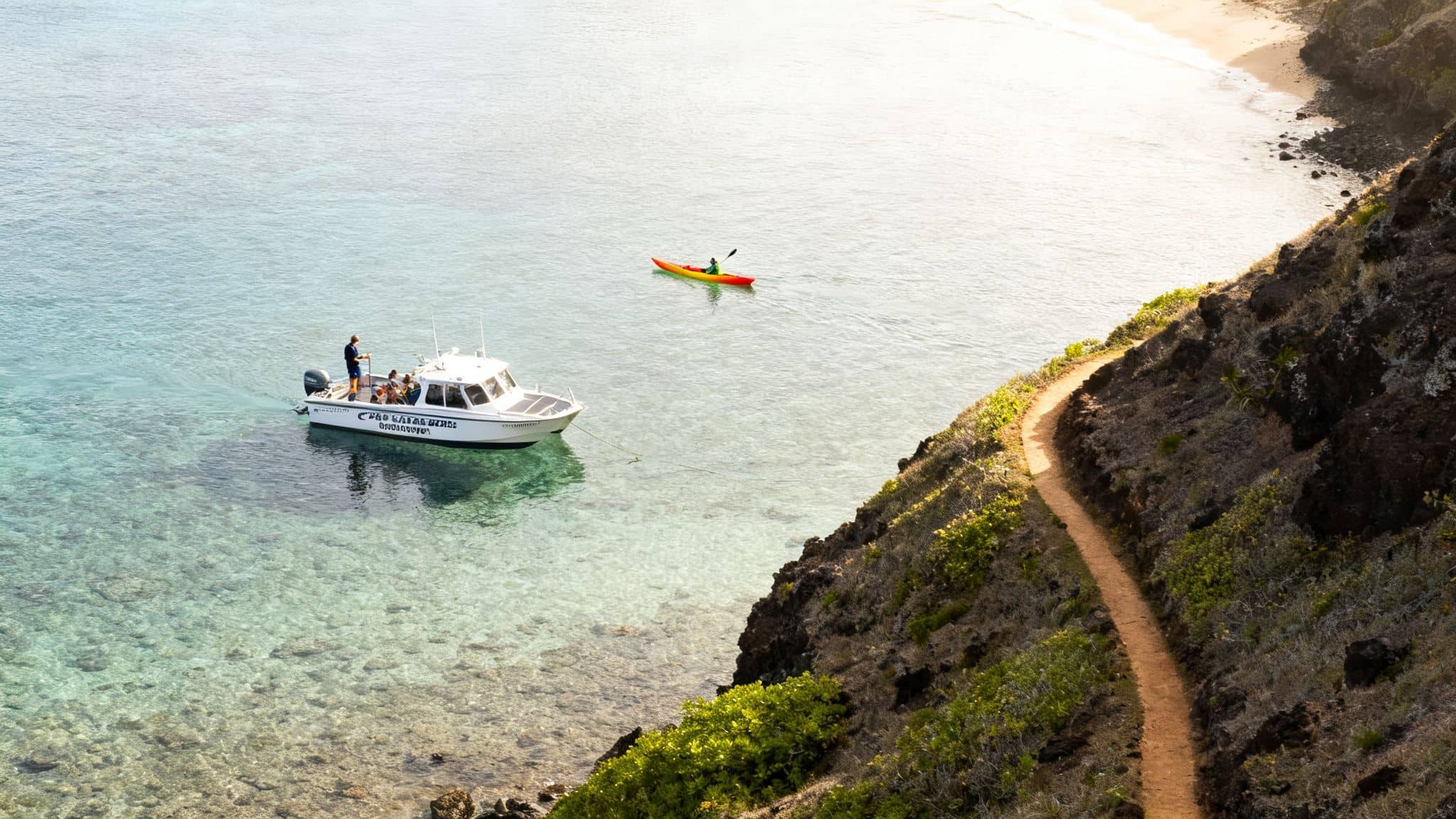 Overhead view of a white boat, a kayak, and a hiking path along a scenic coastline.