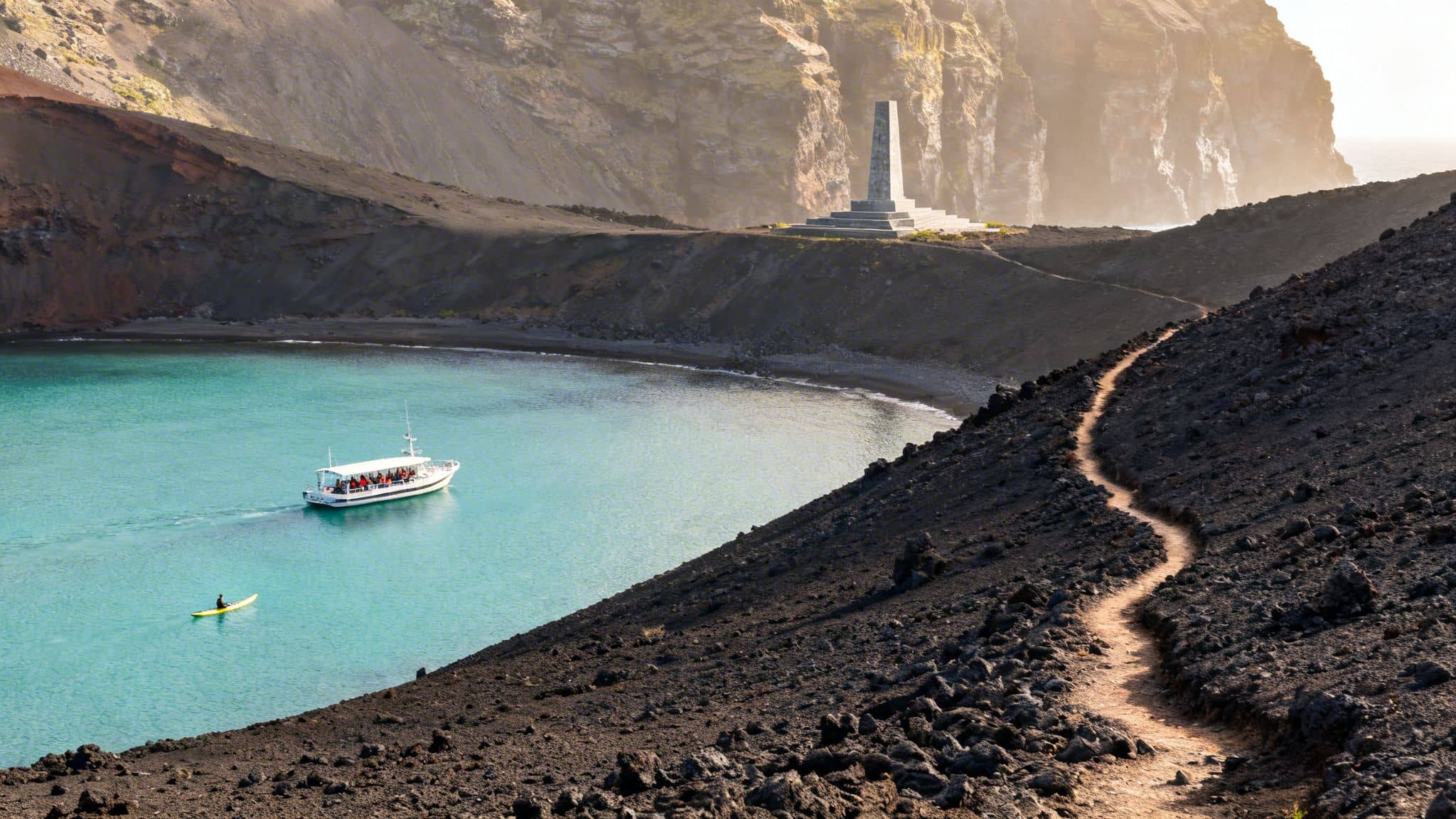 Panoramic view of a bay with turquoise water, a tour boat, kayak, and a monument on volcanic hills.