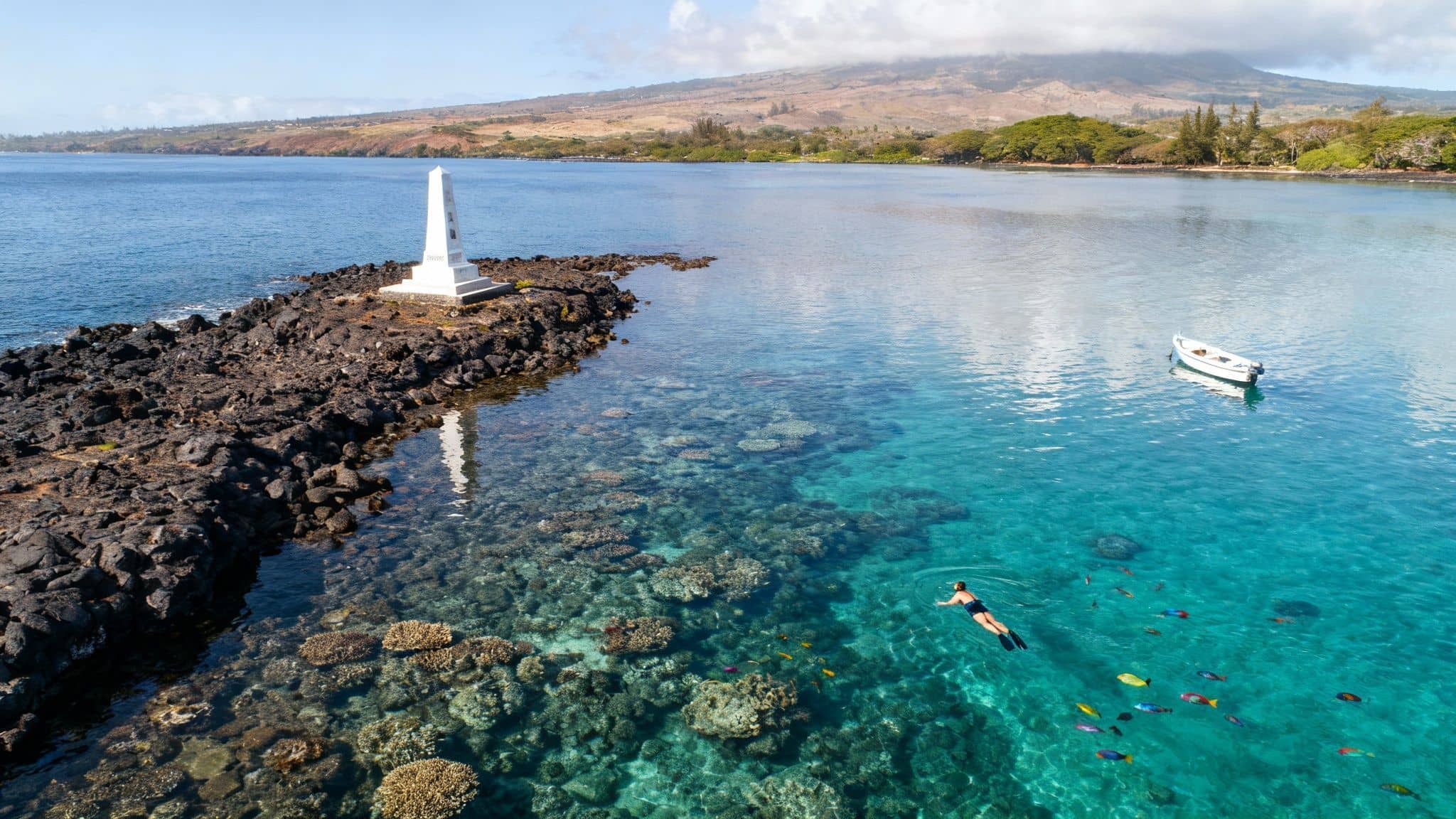 Snorkeler explores vibrant coral reefs and colorful fish near a monument in clear Hawaiian bay.