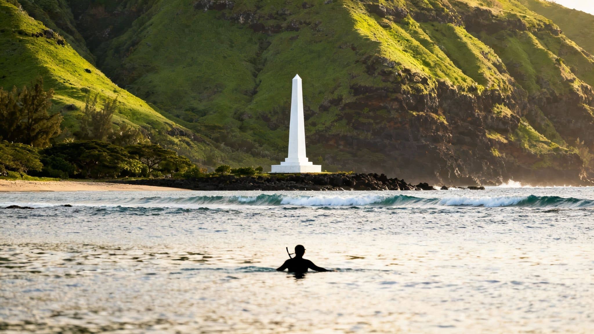 Surfer in ocean near white memorial obelisk with lush green Hawaiian mountains backdrop