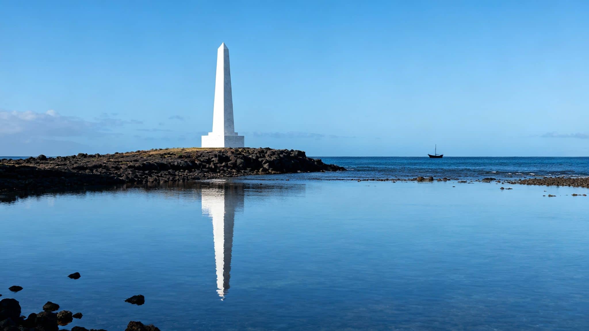 A tall white obelisk stands on a rocky shore, its reflection shimmering in tranquil blue water, with a distant sailboat.