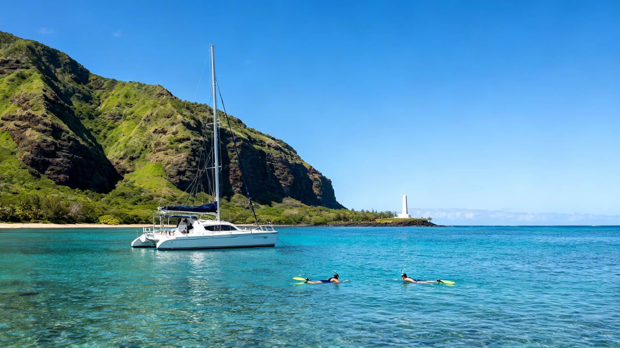 Two people snorkel near a white catamaran in a clear blue bay with a green mountain and lighthouse.