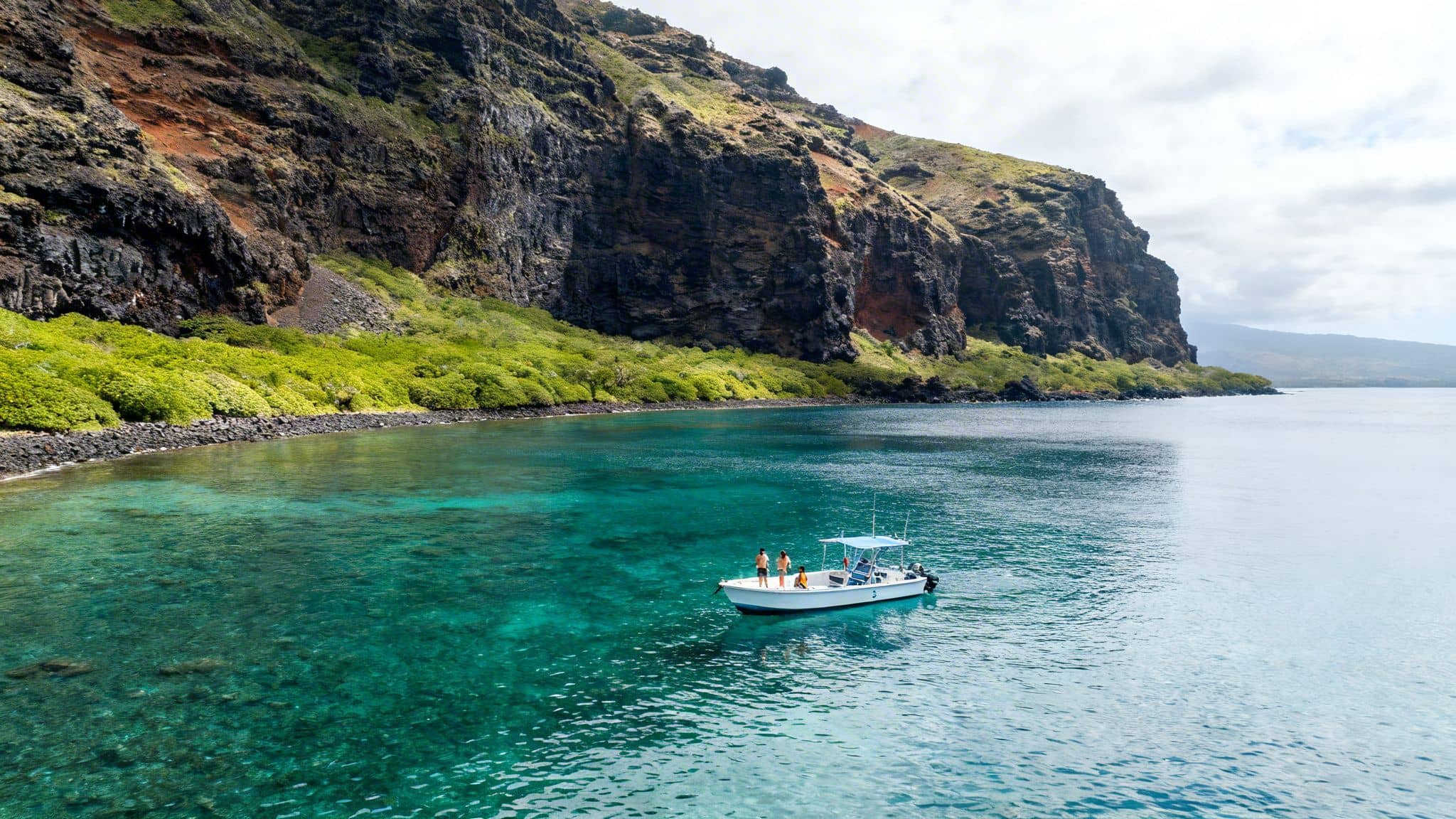 A small boat with people in clear turquoise water near lush green cliffs under a cloudy sky.