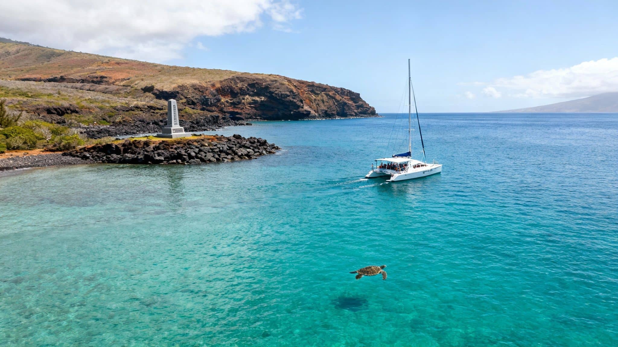 A sea turtle swims in crystal-clear turquoise water near a white catamaran and rocky Hawaiian coastline.