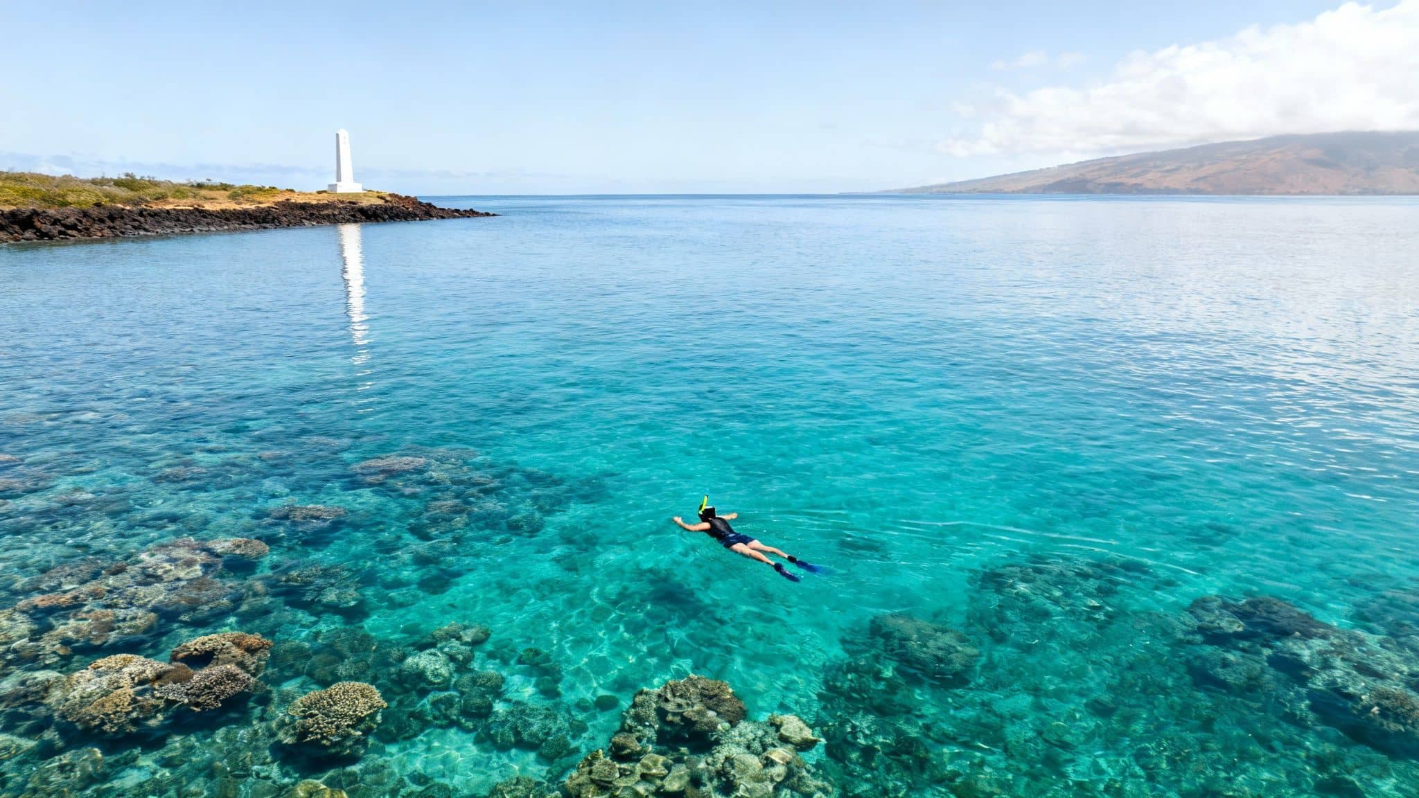 An aerial view of a snorkeler in clear turquoise water over coral reefs near a white lighthouse.