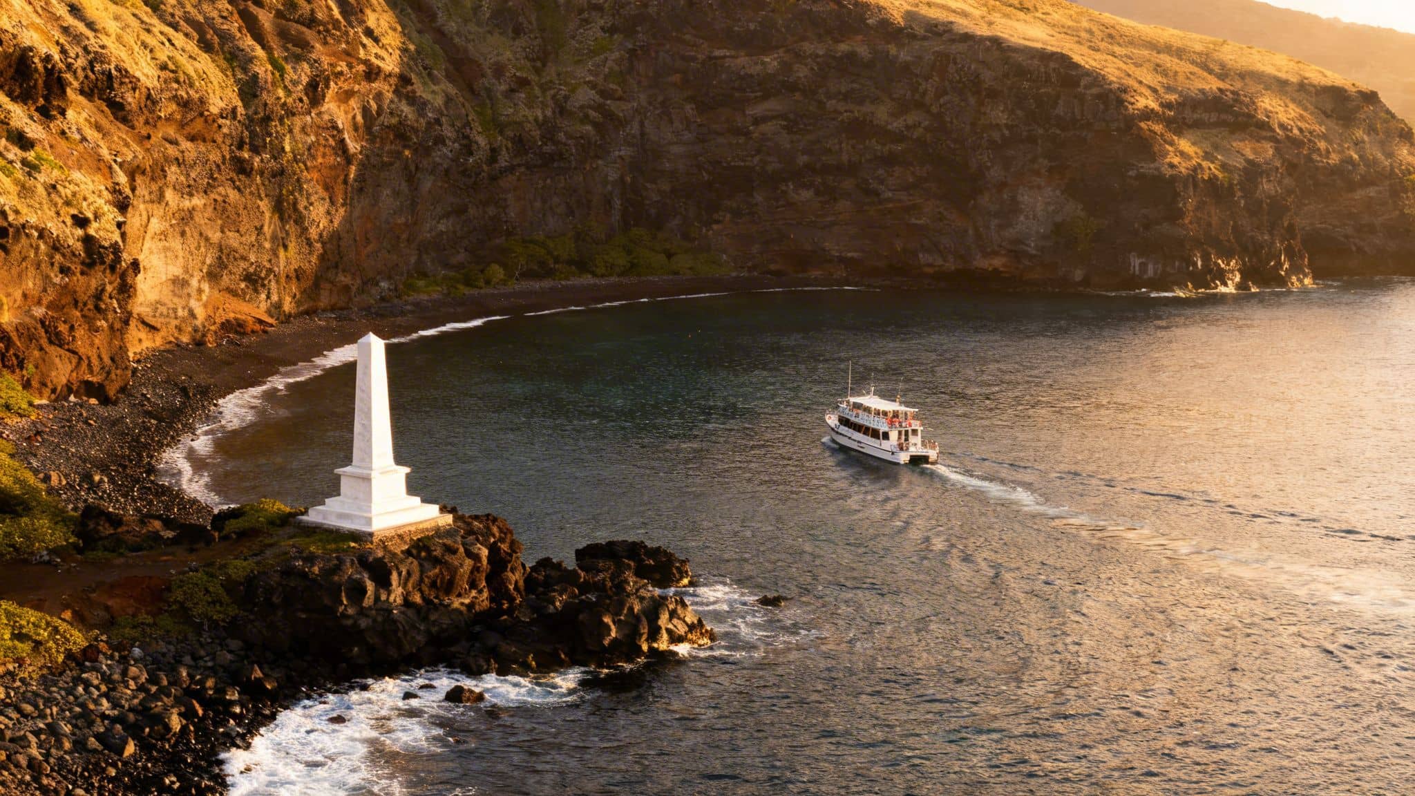 Sunset view of the Captain Cook monument on a rocky point with a tour boat in Kealakekua Bay.