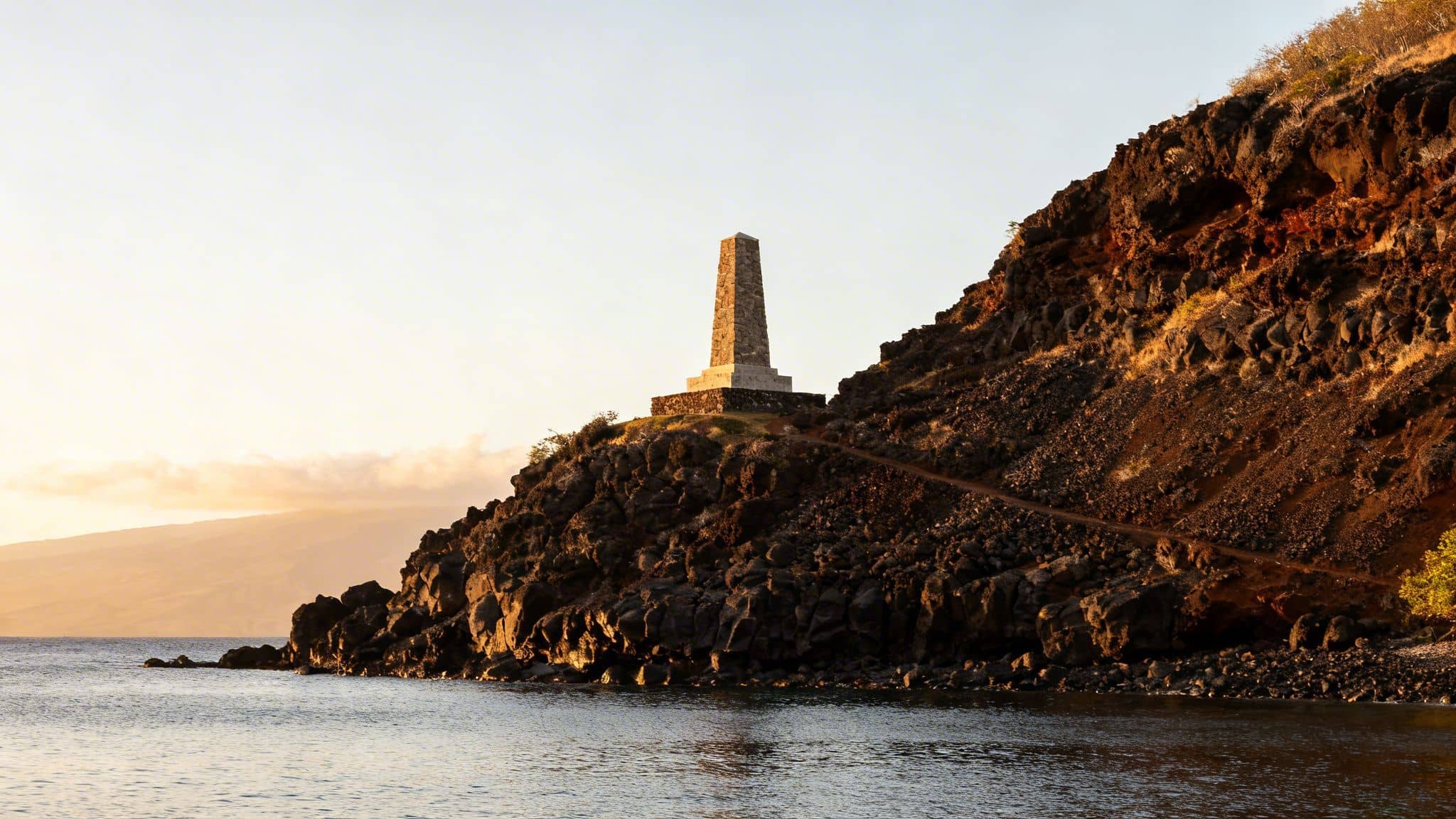 A stone monument on a rocky cliff overlooking the calm ocean at sunrise.